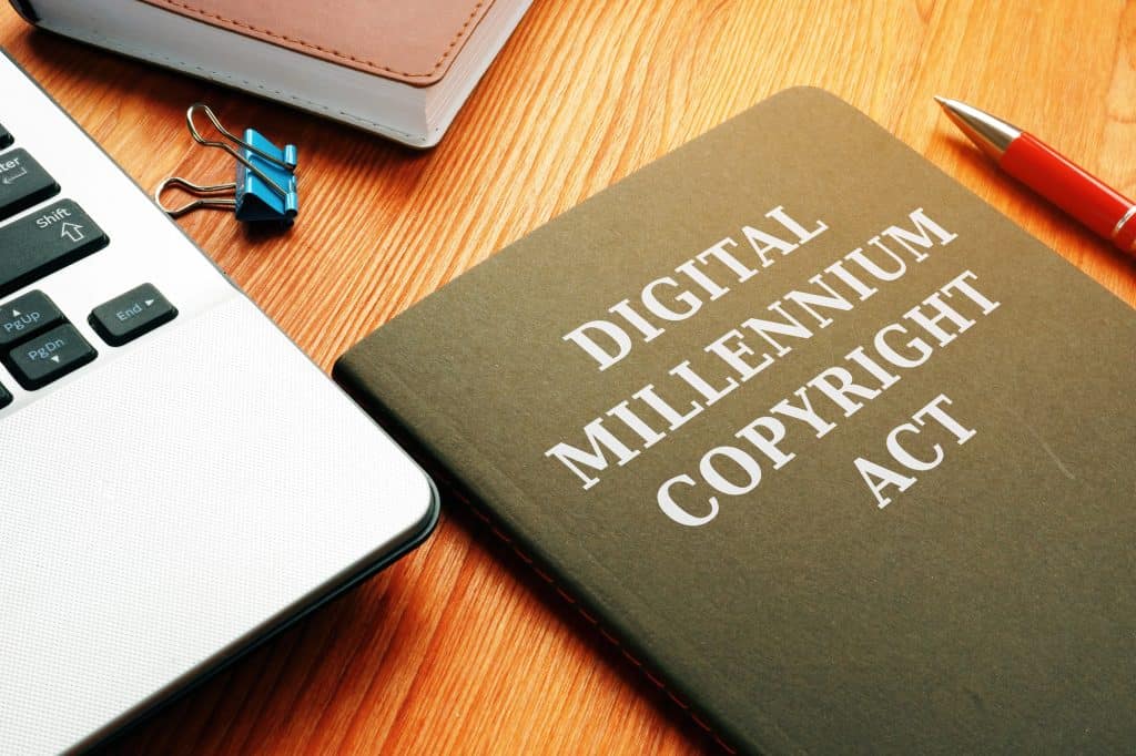 A close-up of a book titled "Digital Millennium Copyright Act" on a wooden surface next to a laptop, notebook, blue binder clip, and a red pen.