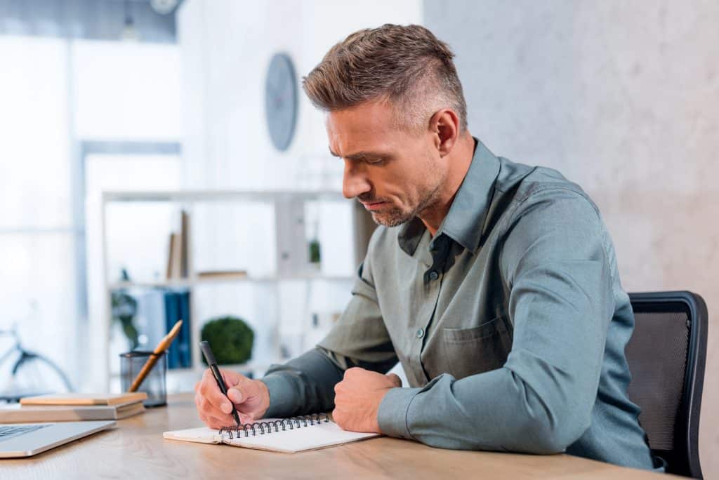 A man in a light gray shirt writing in a notebook at a desk in a bright, modern office.