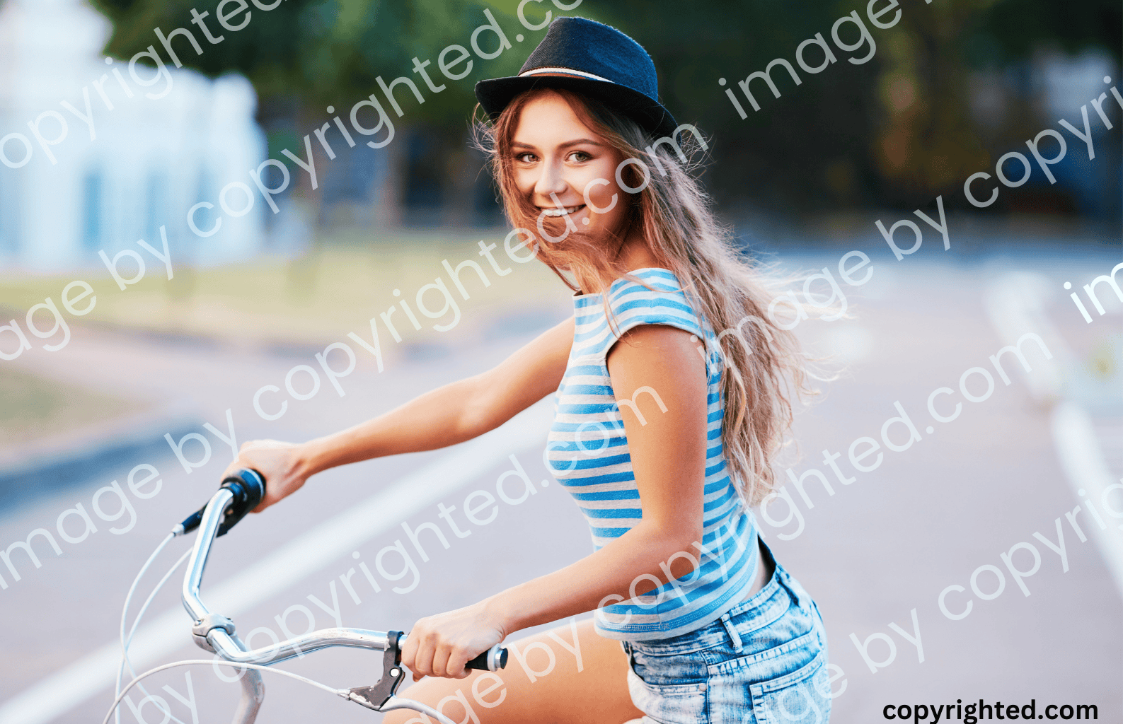 A young woman wearing a striped top and a black hat, smiling while riding a bicycle on an empty road, with a visible watermark across the image.