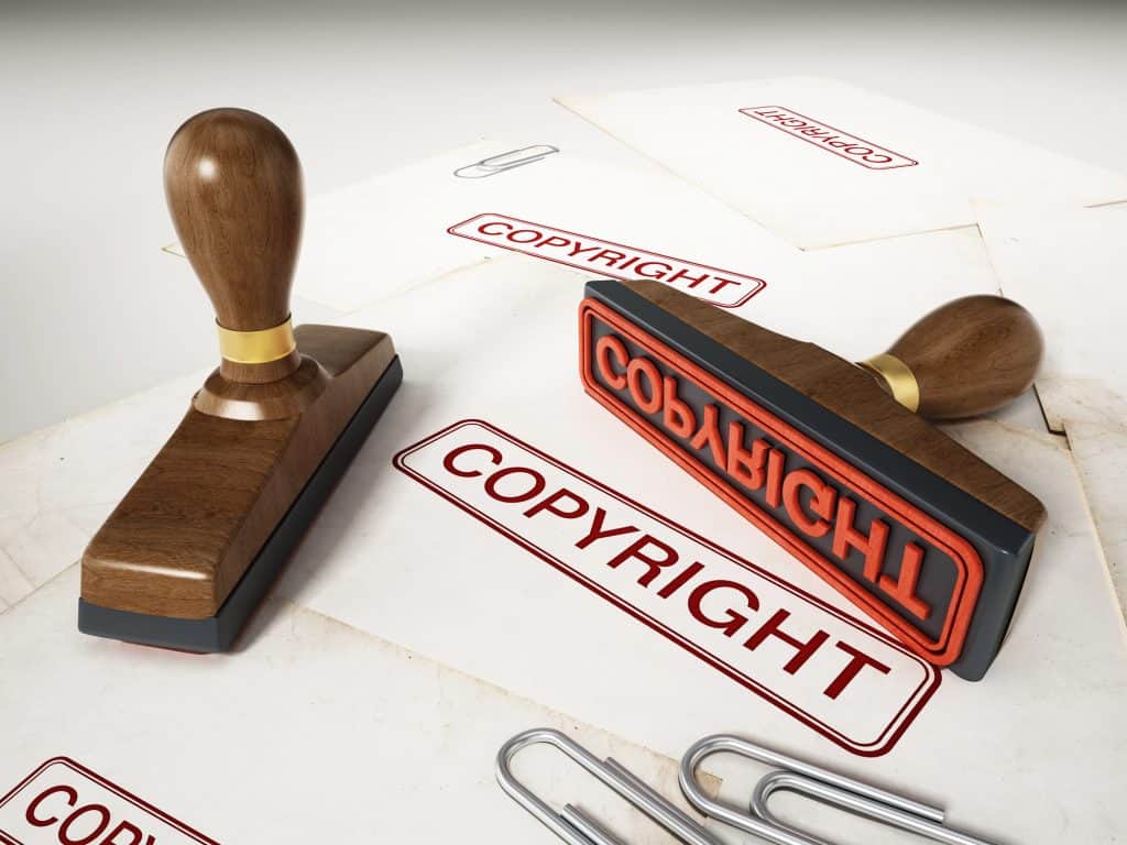 A wooden stamp with the word "COPYRIGHT" is shown on several sheets of paper that have been stamped with the same word, alongside scattered paper clips