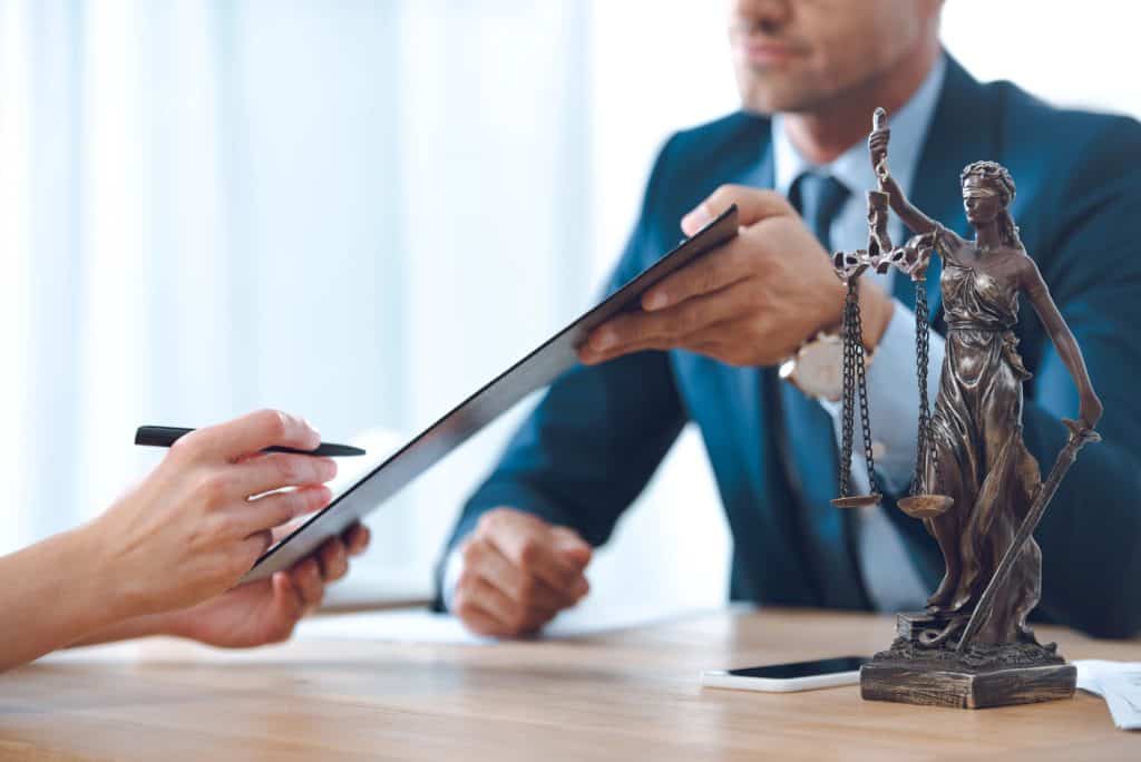 A person handing a clipboard to another person at a desk with a statue of Lady Justice in the foreground.