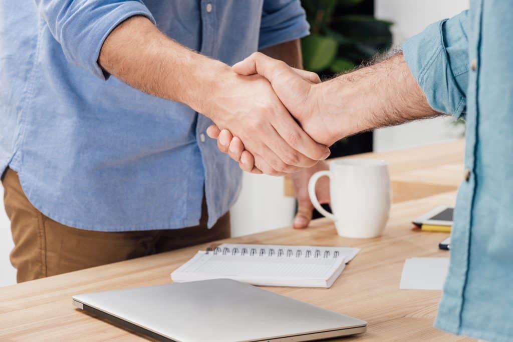 Two people shaking hands across a desk with a laptop, notebook, and coffee cup in the background.