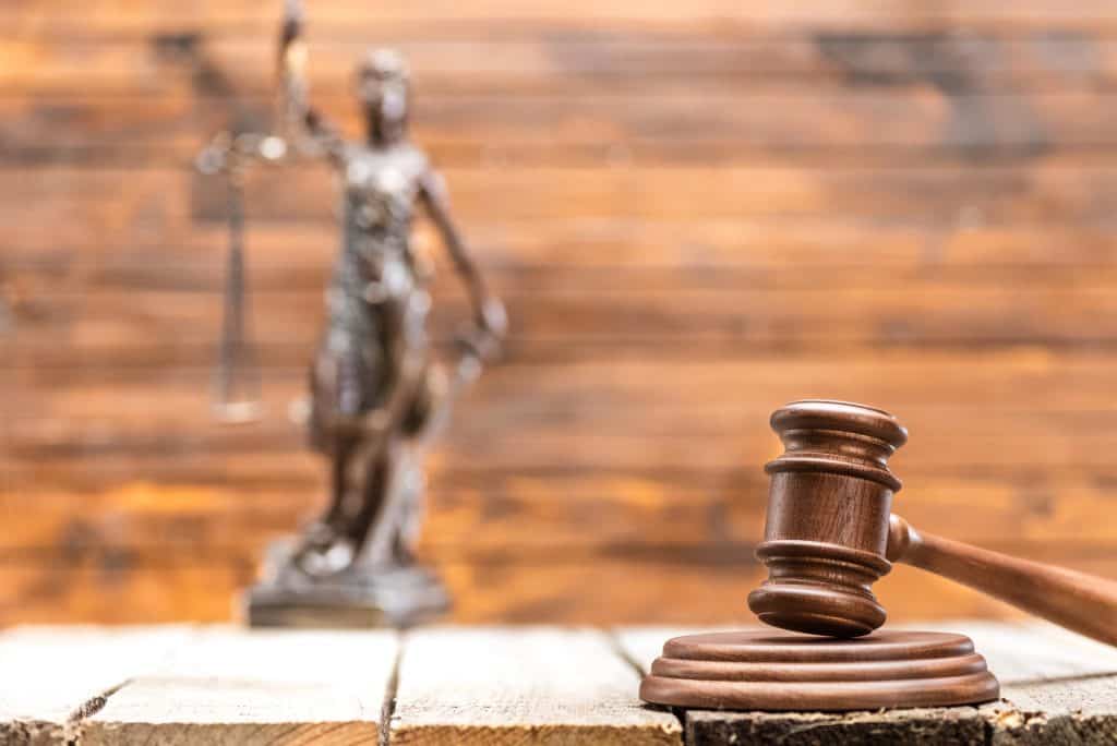 A wooden gavel on a table with a blurred statue of Lady Justice holding scales in the background.