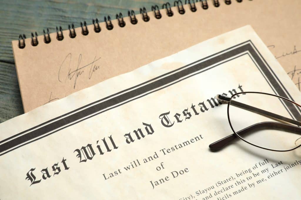 A last will and testament document with glasses resting on it and a signed notebook in the background.