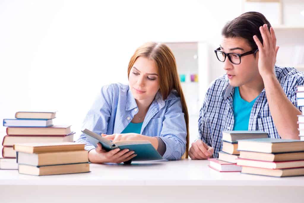 A male and female student surrounded by stacks of books, with the female pointing to a page and the male looking surprised or confused.