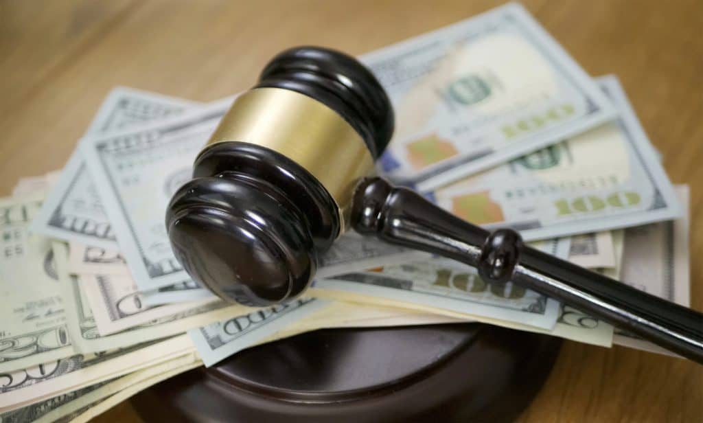 A wooden gavel resting on a pile of hundred-dollar bills on a wooden surface.