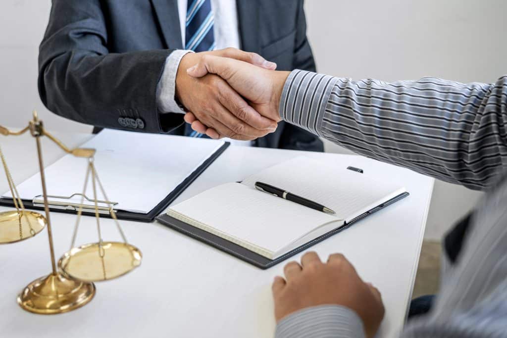 Two individuals shaking hands across a desk with legal documents, a pen, and a brass balance scale on the table.