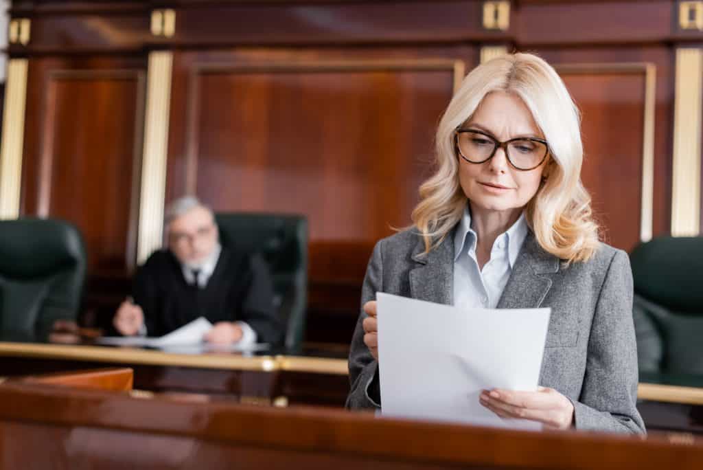 A female lawyer reading a document in a courtroom, with a judge seated in the background.