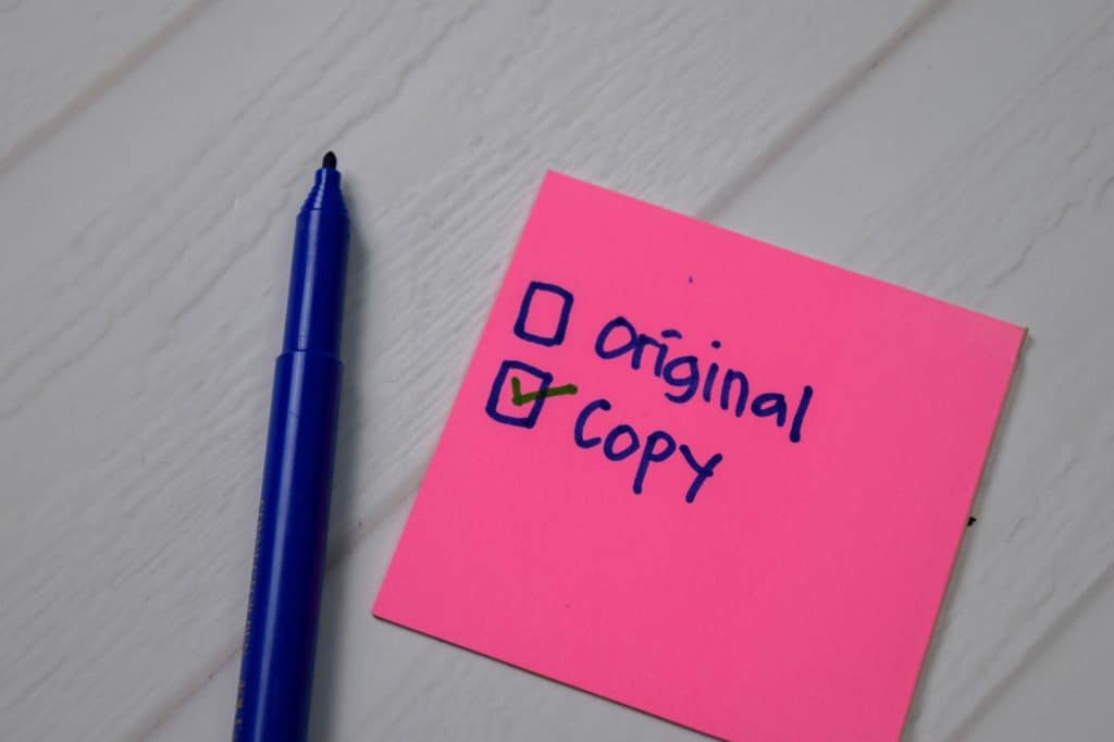 A pink sticky note with a checklist, where "copy" is checked, next to a blue marker on a white surface.