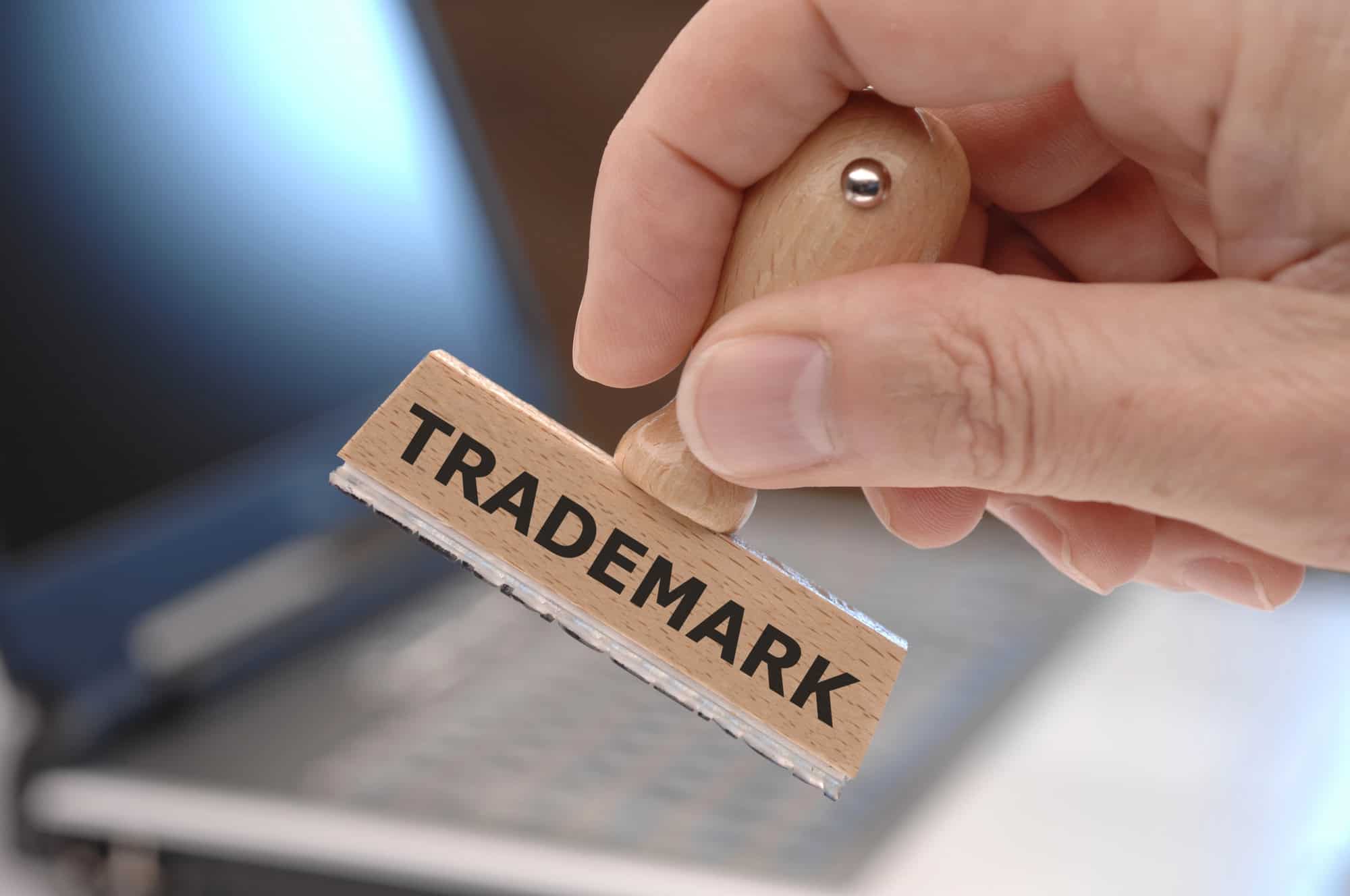 A hand holding a wooden stamp with the word "Trademark" over a blurred background.