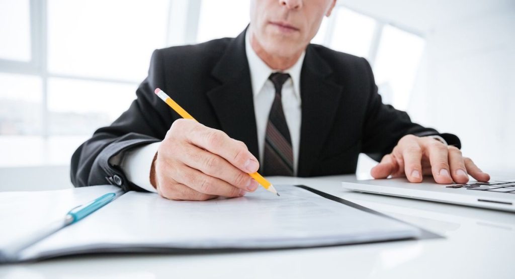 A man in a suit is sitting at a desk, writing on a document with a pencil.