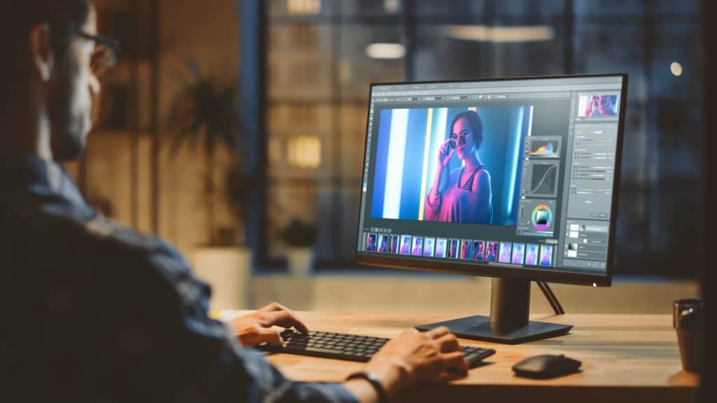 A person sitting at a desk using photo editing software on a desktop monitor, with an image of a woman wearing sunglasses displayed on the screen.