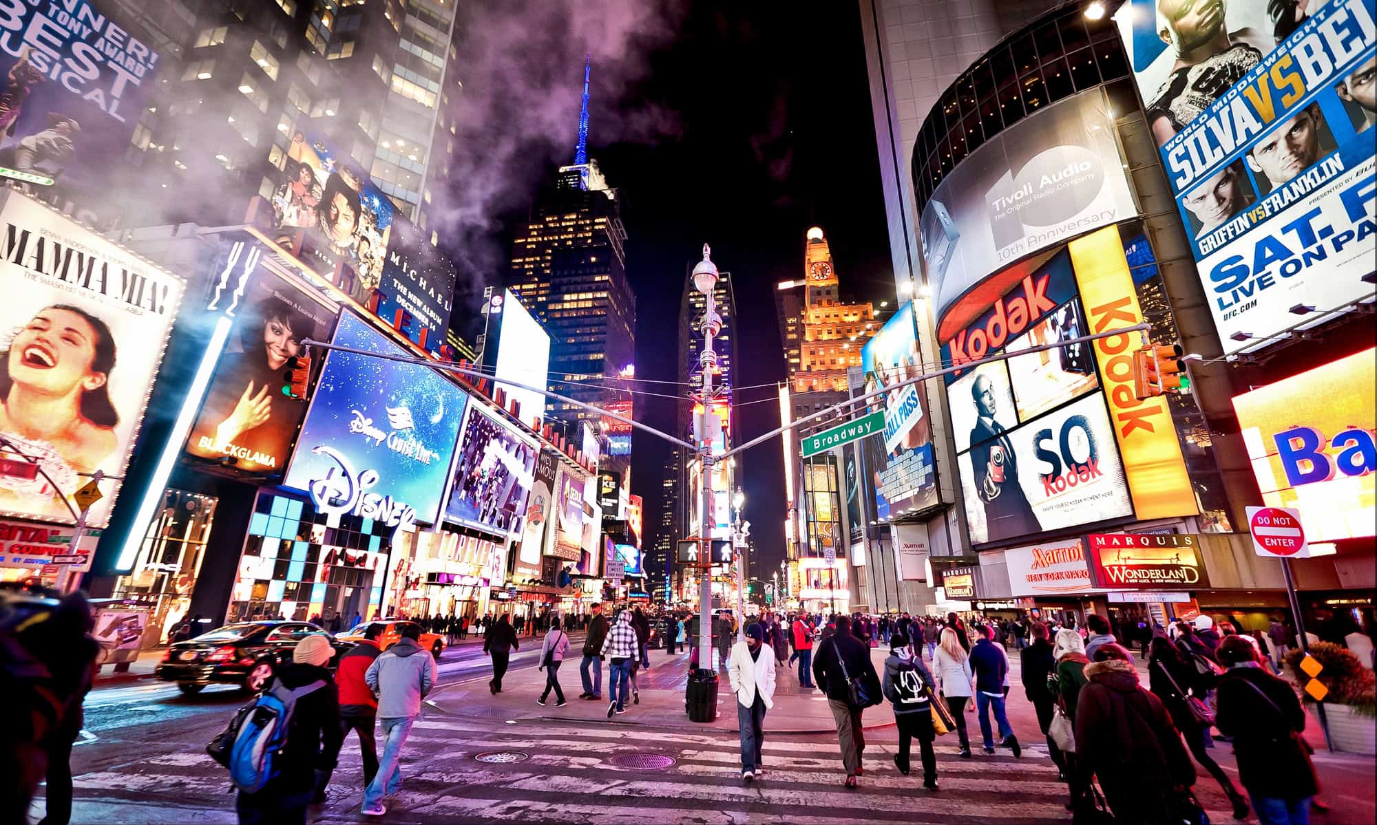 A vibrant nighttime scene of Times Square in New York City, filled with bright billboards, neon signs, a crowd of pedestrians, and surrounding skyscrapers.